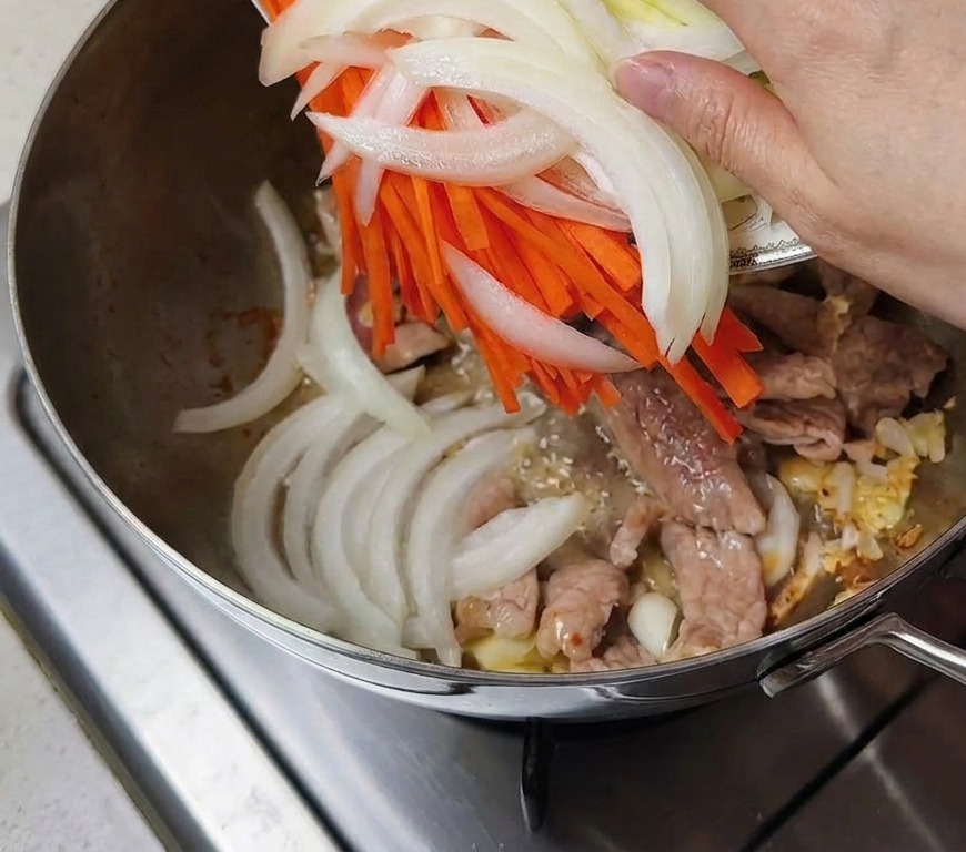 Zwiebeln, Karotten und Zucchini werden zu dem Fleisch in den Topf gegeben.