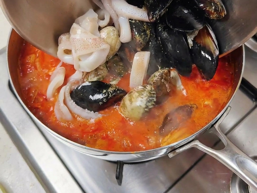 Muscheln und Tintenfischringe werden aus einer Schüssel in die kochende rote Brühe gegeben.