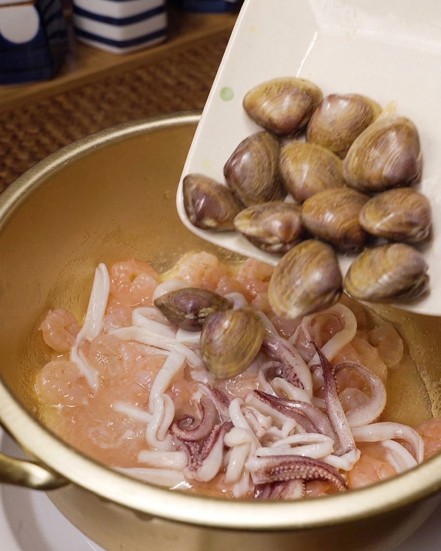 Frische Muscheln werden von einem weißen Teller in einen Topf zu bereits angebratenen Garnelen und Tintenfischringen gegeben.