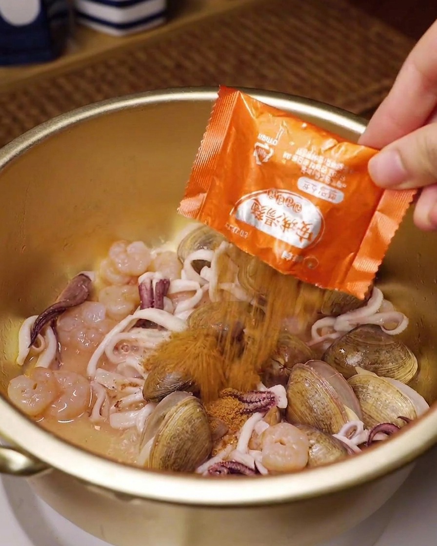 Eine Hand schüttet ein Päckchen Instant-Ramen-Gewürz über Meeresfrüchte in einem goldenen Kochtopf.