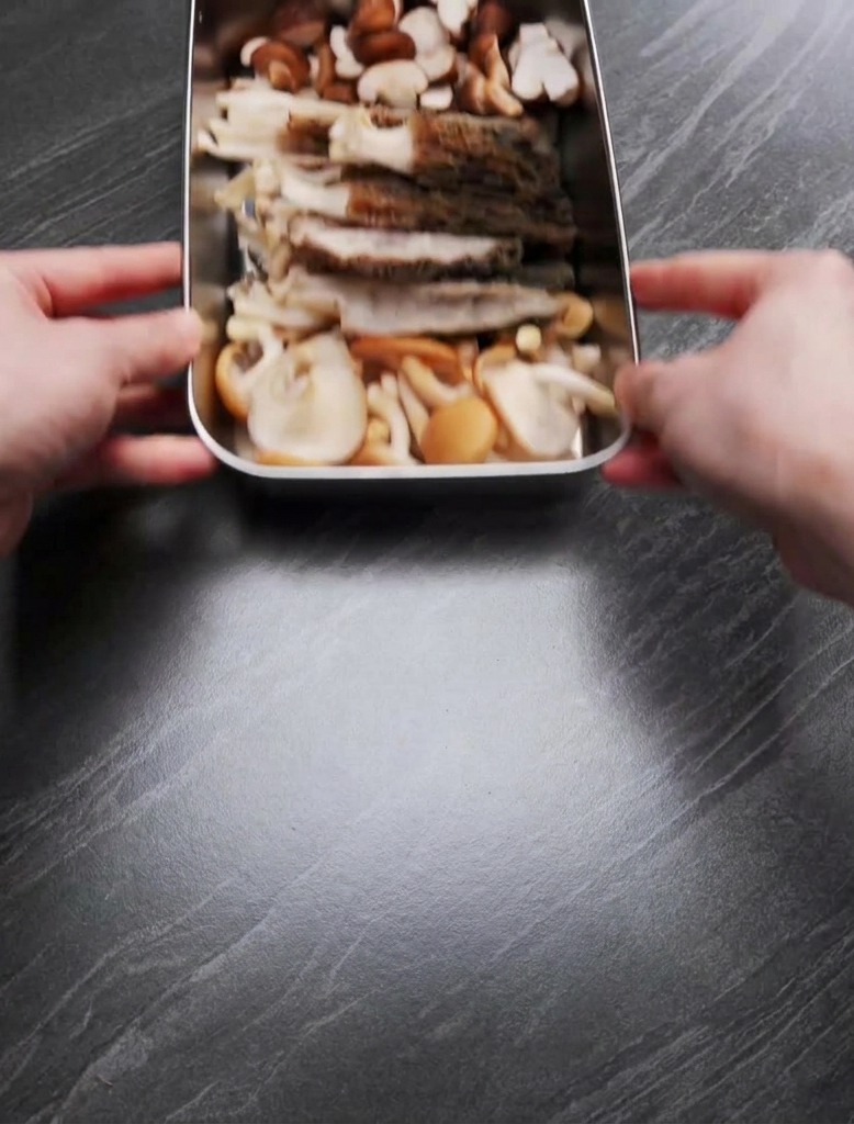 Hände halten ein Tablett mit geviertelten Morcheln und halbierten Shiitake-Pilzen auf einer dunklen Arbeitsplatte.