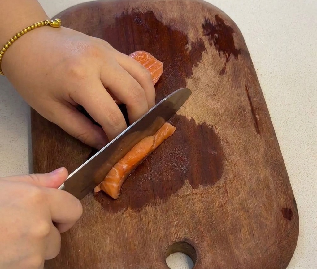 Eine Hand schneidet ein frisches, orangefarbenes Lachsfilet auf einem dunklen Holzbrett in gleichmäßige kleine Würfel.