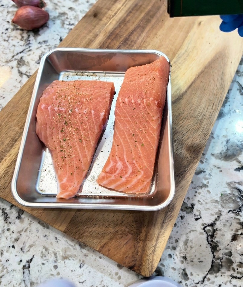 Eine Hand würzt rohe Lachsfilets in einer Schale mit einer Prise schwarzem Pfeffer und Knoblauchpulver.