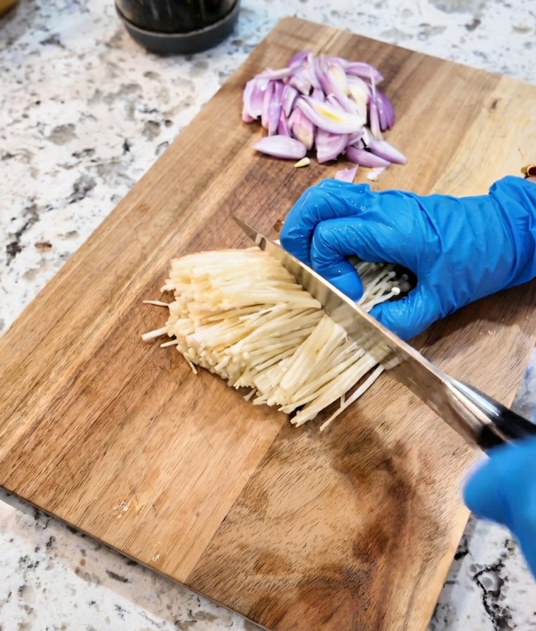 Ein Messer schneidet die Wurzelenden eines Bundes weißer Enoki-Pilze auf einem Schneidebrett ab.