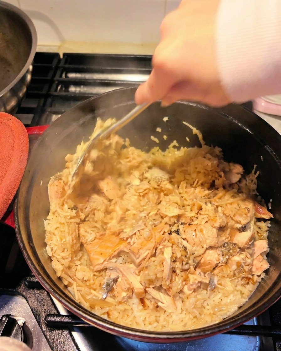 Eine Hand zerteilt mit einem Metallwender den zarten Lachs und mischt ihn unter den Reis im Topf.