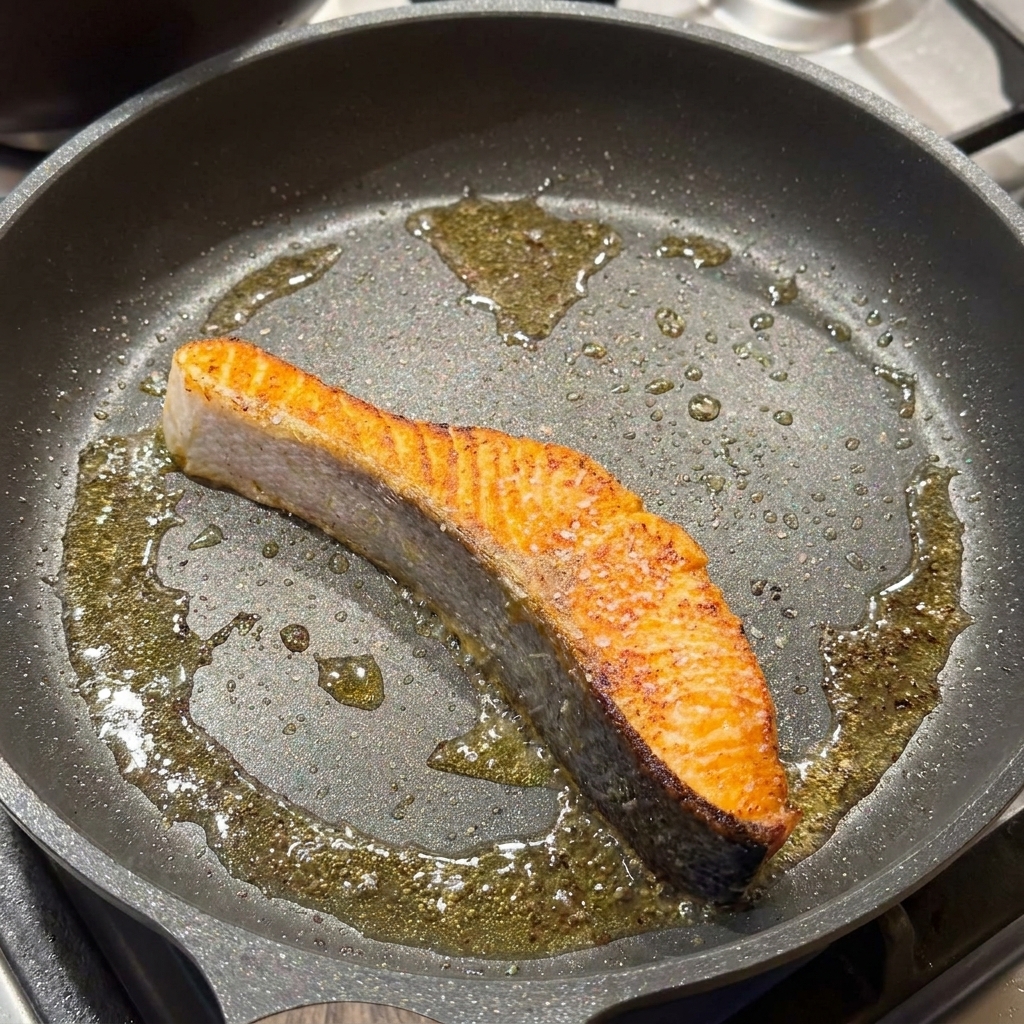 Ein angebratenes Lachsfilet in einer Bratpfanne wird mit Salz und schwarzem Pfeffer gewürzt.
