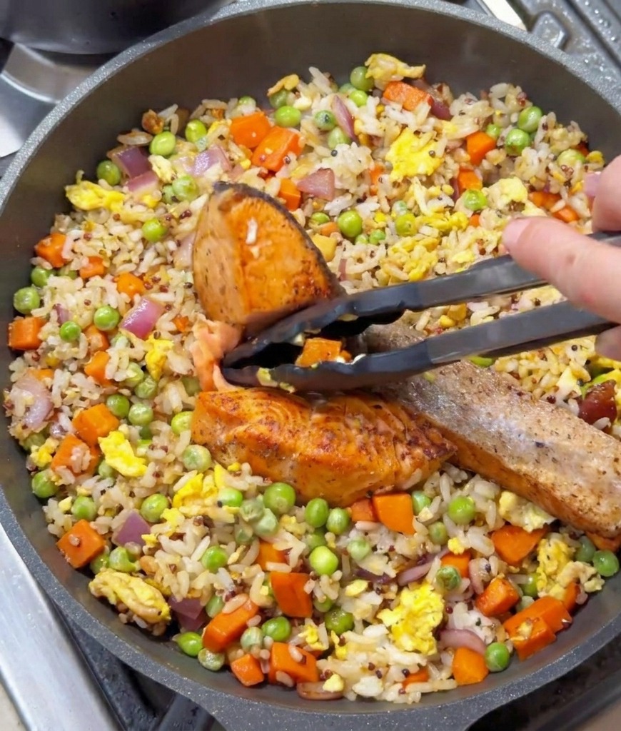 Eine schwarze Küchenzange hält ein Stück gebratenen Lachs über eine Pfanne mit Bratreis und Gemüse.