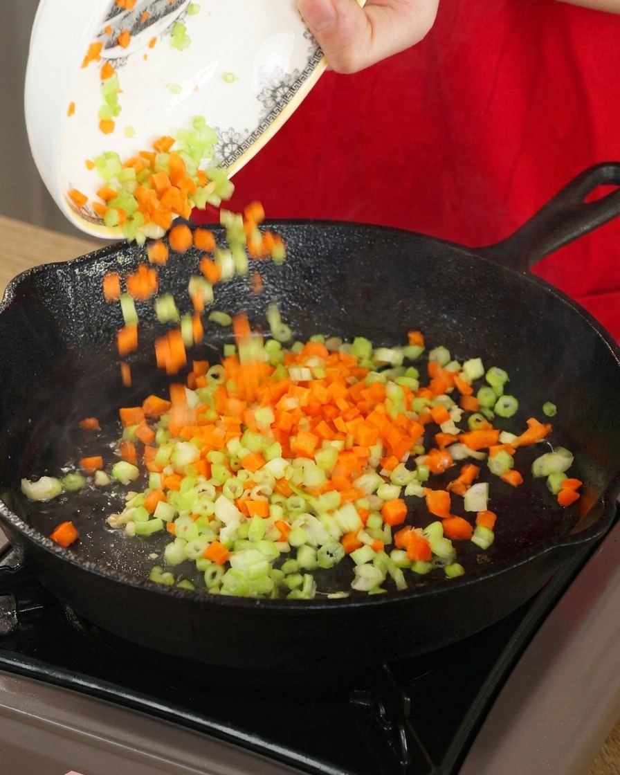 Gewürfelte orangefarbene Karotten und grüner Sellerie werden aus einer weißen Schüssel in eine heiße, schwarze Gusseisenpfanne geschüttet.