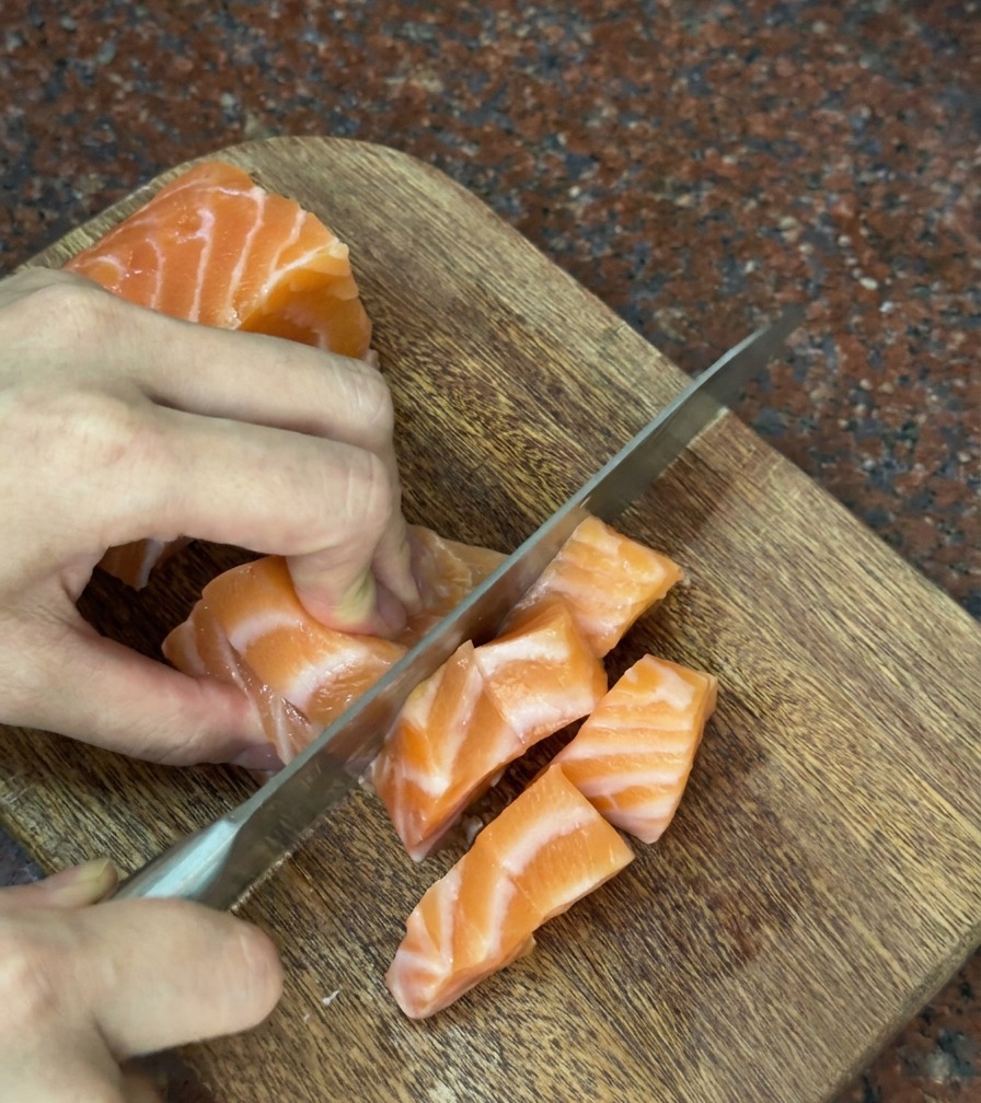 Hände schneiden vorsichtig ein frisches Stück orangefarbenen Lachs in dicke, quadratische Würfel auf einem Holzbrett.