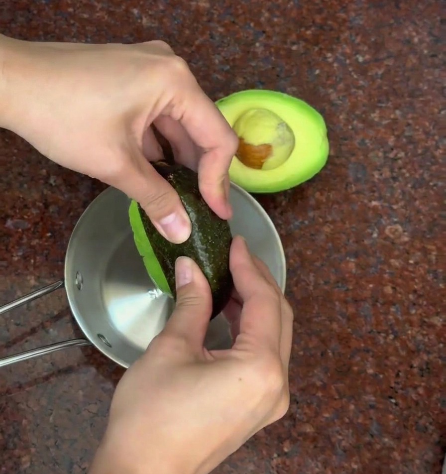 Eine Person zieht mit den Händen die dunkle Schale von einer halbierten Avocado über einer Metallschüssel ab.