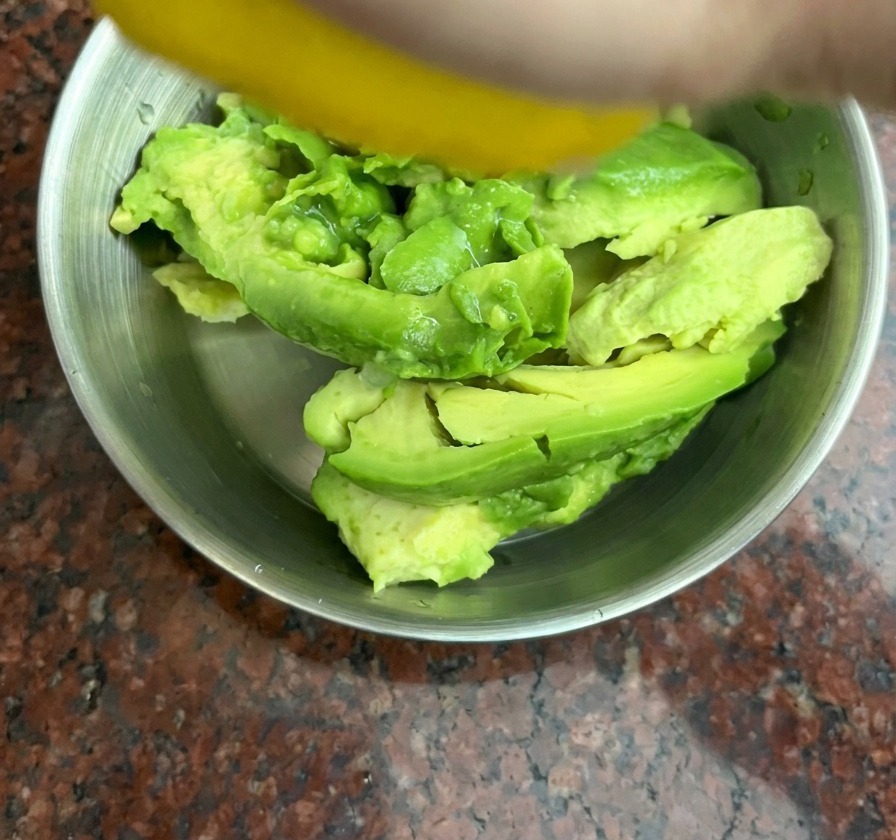 Eine Hand presst eine gelbe Zitrone über Avocado-Stücken in einer Edelstahlschüssel aus.