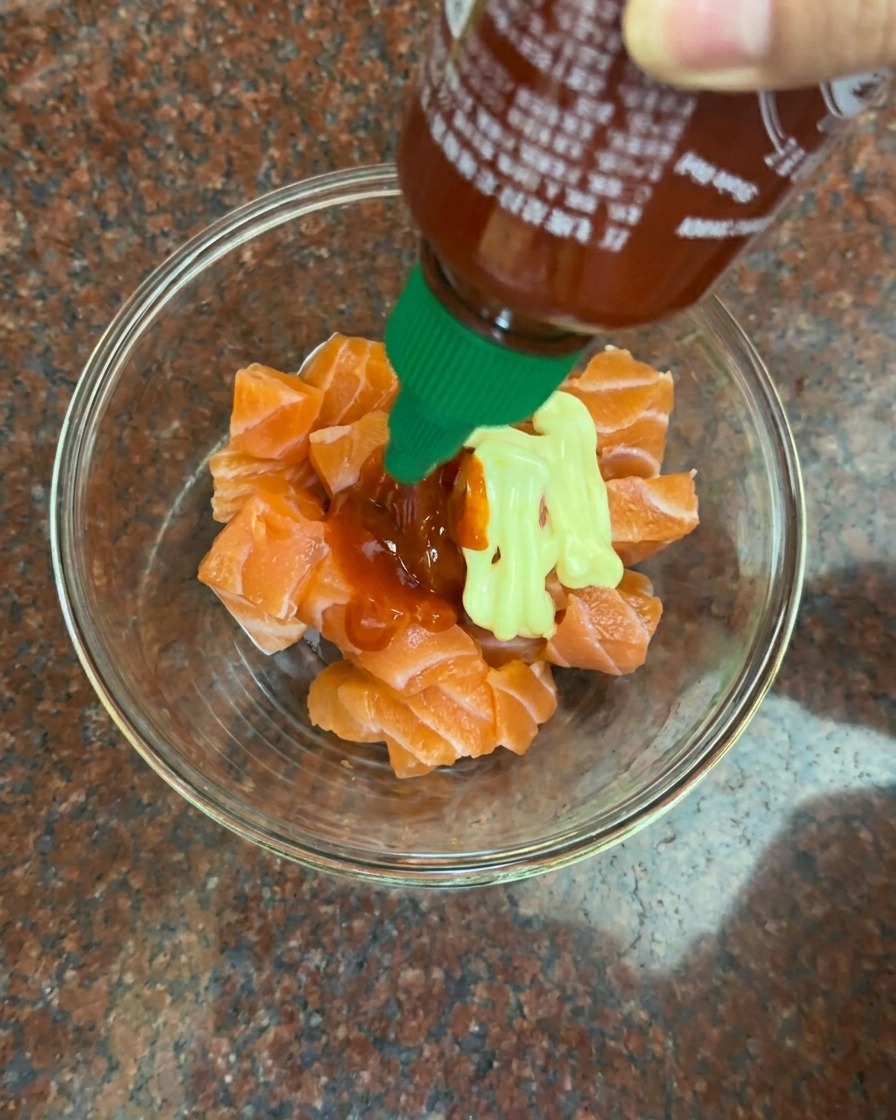 Cremige Mayonnaise und leuchtend rote Chilisauce werden zu einer Schüssel mit rohen Lachswürfeln gegeben.