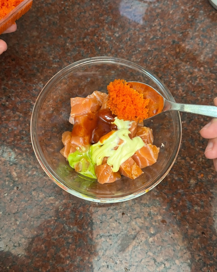 Ein Löffel gibt eine große Portion winzigen orangen Fischrogen in eine Schüssel mit Lachs, Mayonnaise und Chilisauce.