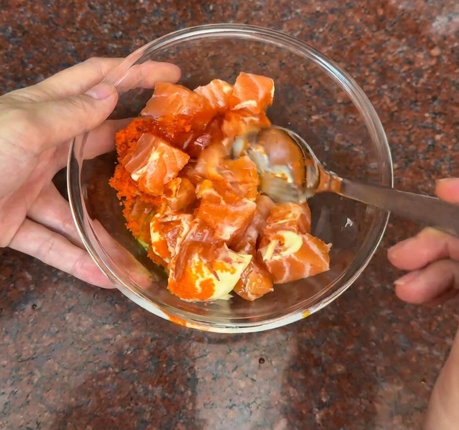 Eine Glasschüssel mit frischen Lachswürfeln, die mit orangem Fischrogen und einer cremigen orangefarbenen Sauce vermengt werden.