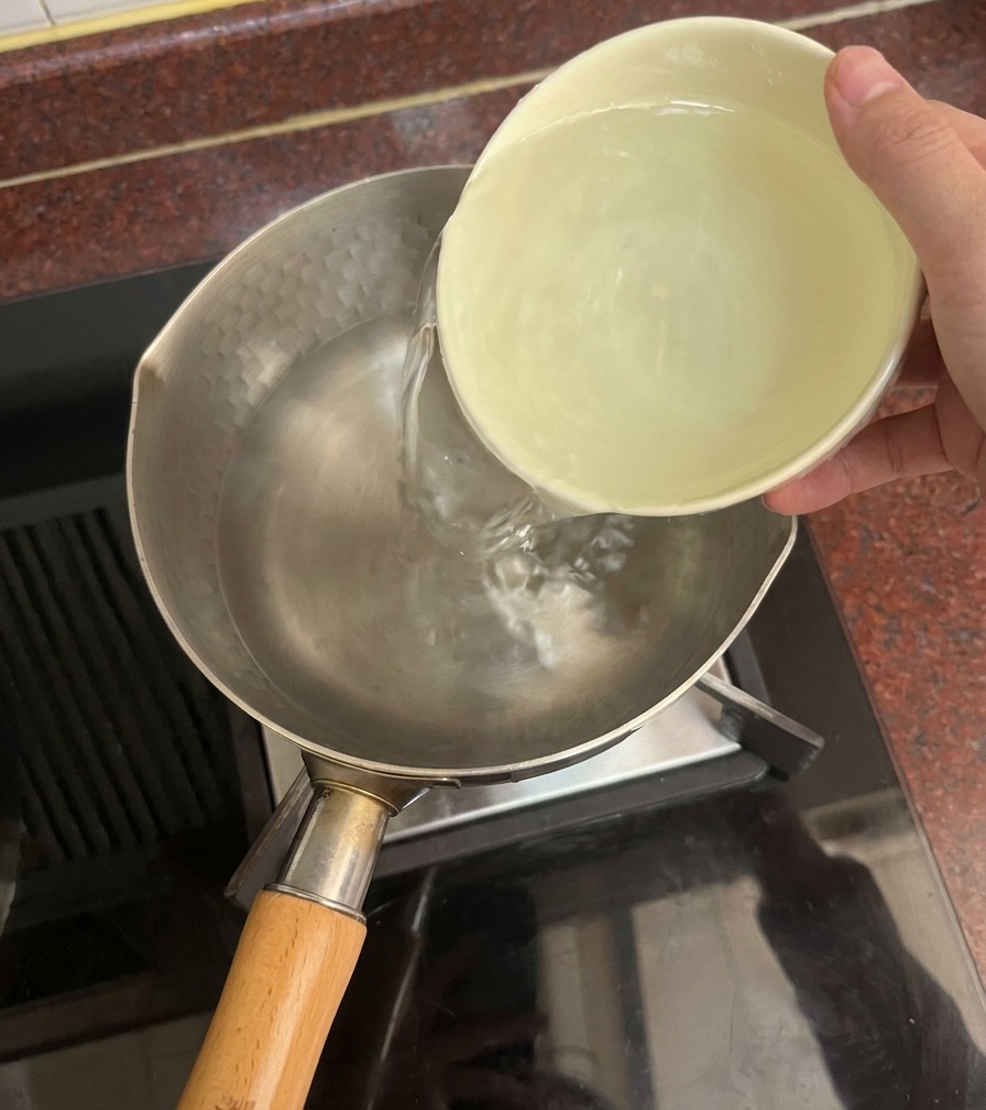Eine Hand gießt klares Wasser aus einer hellen Keramikschüssel in einen Metalltopf auf dem Herd.
