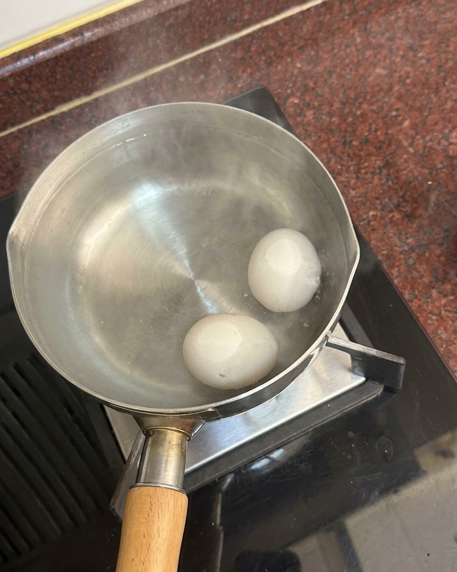 Zwei weiße Eier liegen am Boden eines Topfes mit heißem Wasser, von der Oberfläche steigt Dampf auf.