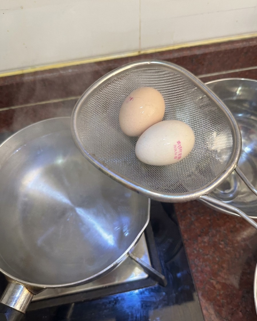 Ein Metallsieb hält zwei Eier über einem Topf, bereit, sie in eine Schüssel mit Eiswasser zu legen.
