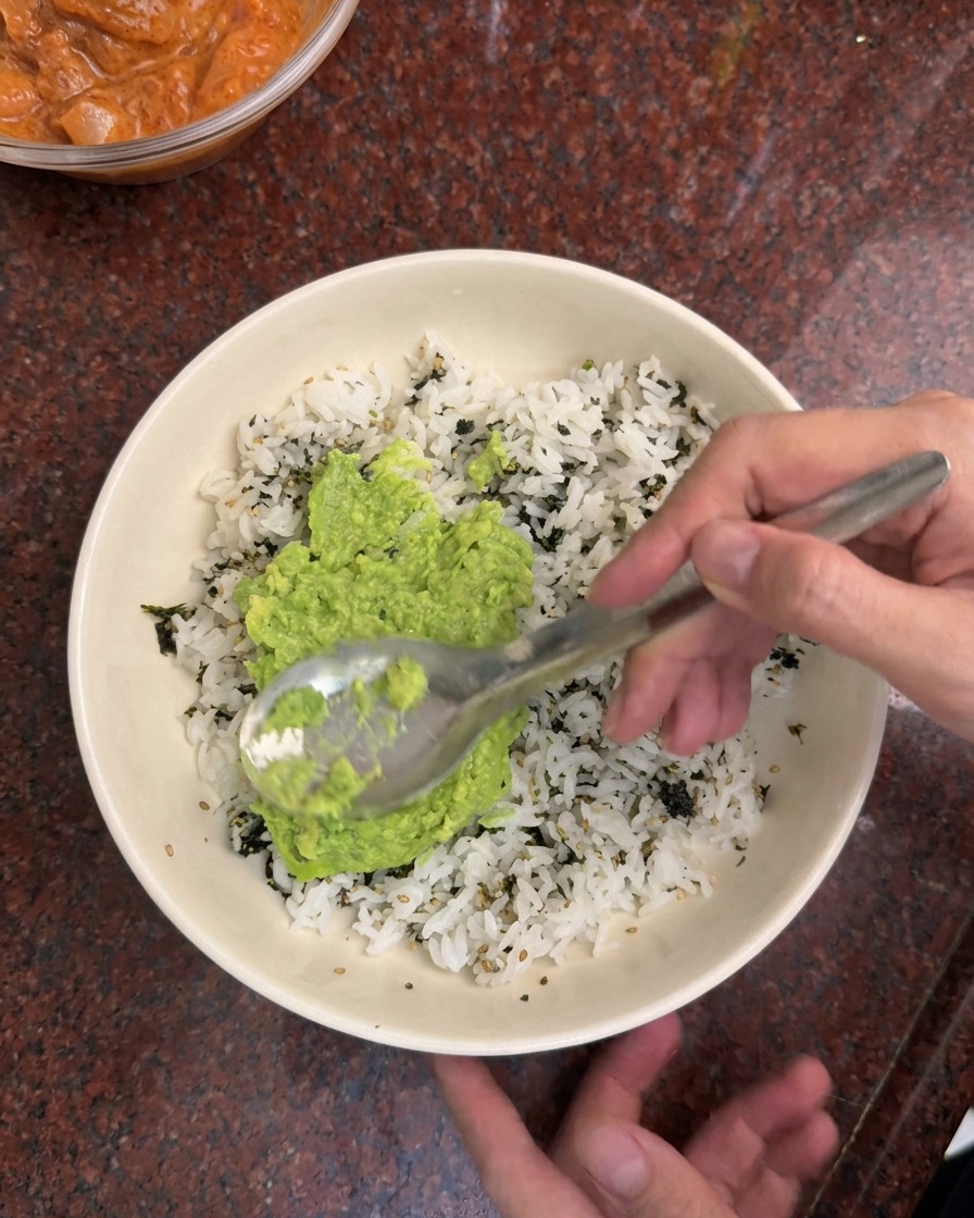 Ein Löffel verteilt hellgrüne Avocado-Creme auf weißem Reis und dunklen Algenflocken in einer weißen Schüssel.