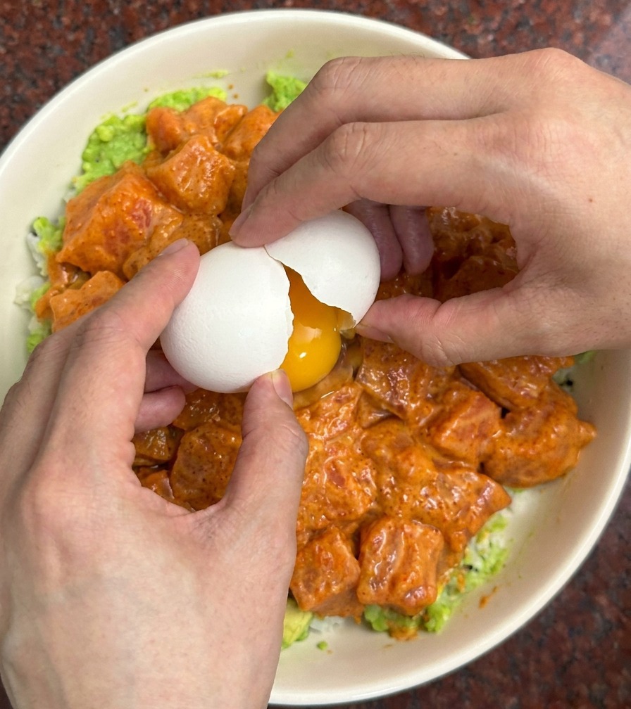 Zwei Hände schlagen eine weiße Eierschale über einer farbenfrohen Bowl mit mariniertem Lachs und Avocado auf.