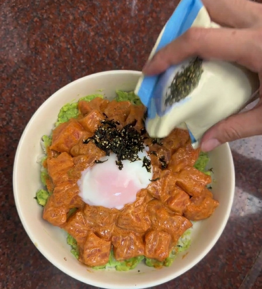 Eine Hand streut eine Mischung aus schwarzen Algenstreifen und Sesam aus einem Beutel auf die Lachs-Bowl.