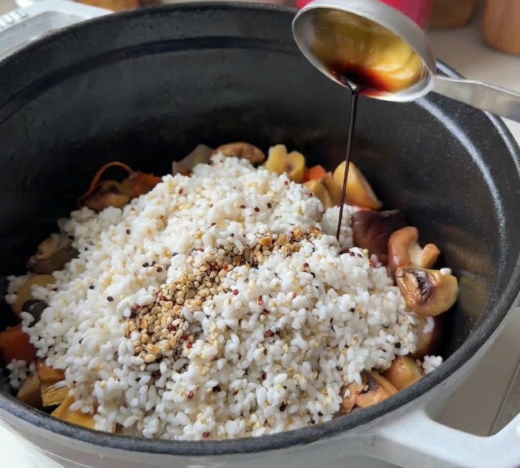 Ein Metalllöffel gießt dunkle Sojasauce über Reis, Quinoa und Pilze in einem schwarzen Topf.