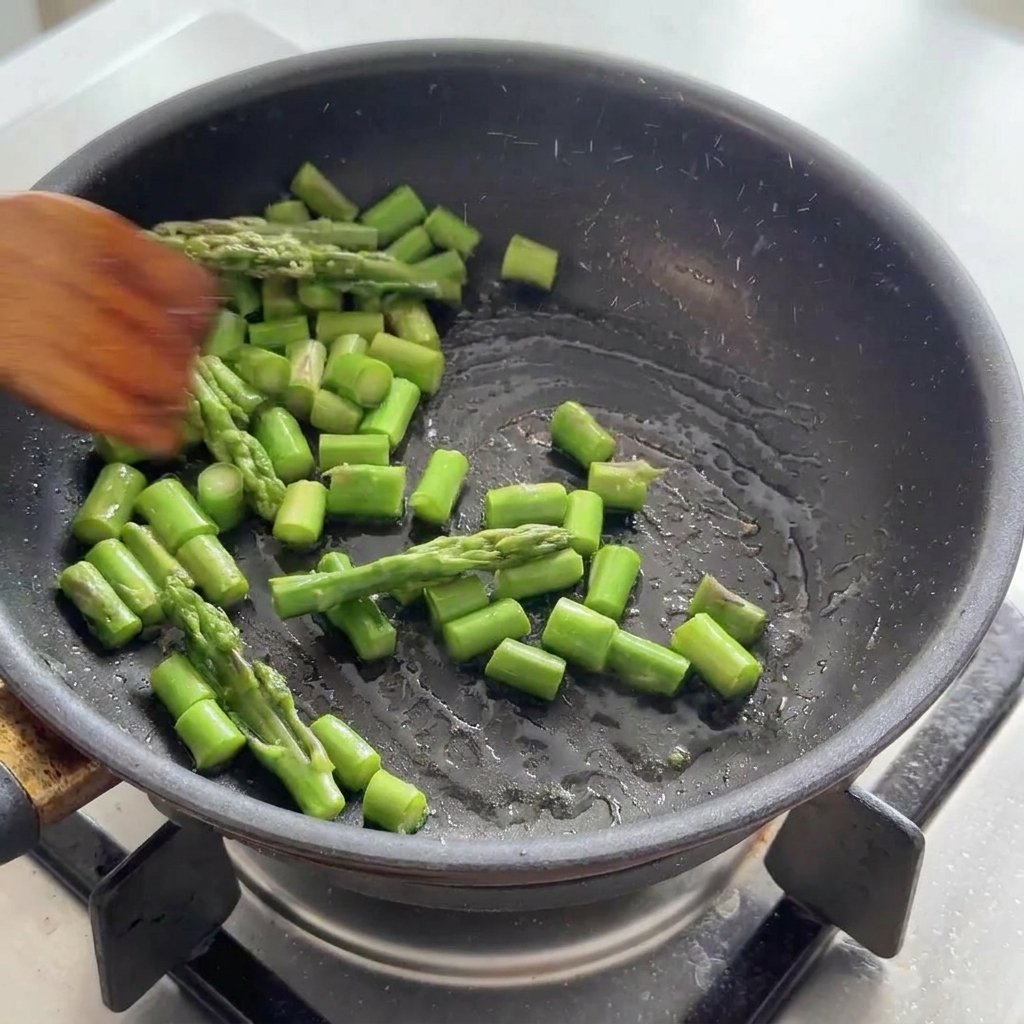Grüne Spargelstücke werden mit einem Holzspatel in einer schwarzen Pfanne geschwenkt.