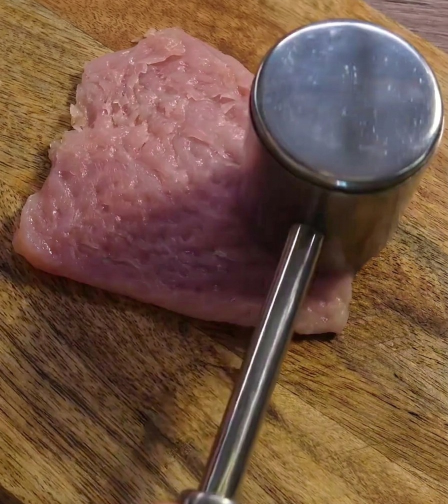 Ein Fleischklopfer bearbeitet eine Scheibe Schweinefleisch auf einem Holzbrett.