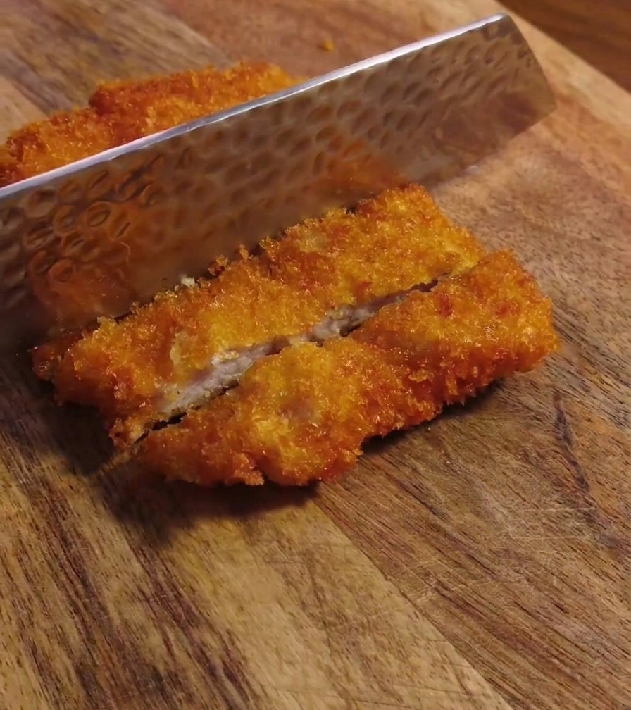 Ein Messer schneidet ein knuspriges Schweineschnitzel auf einem Holzbrett in Streifen.