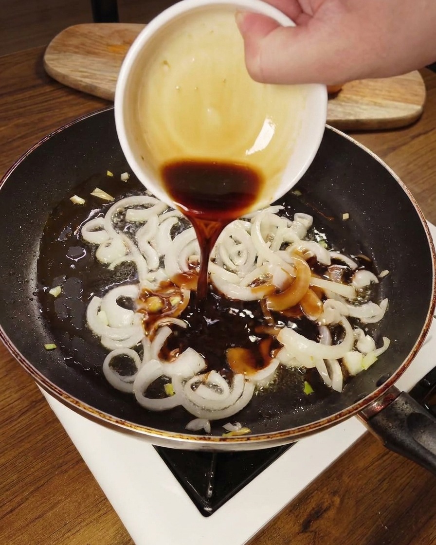 Eine dunkle Würzsauce wird über die gedünsteten Zwiebeln in der Pfanne gegossen.