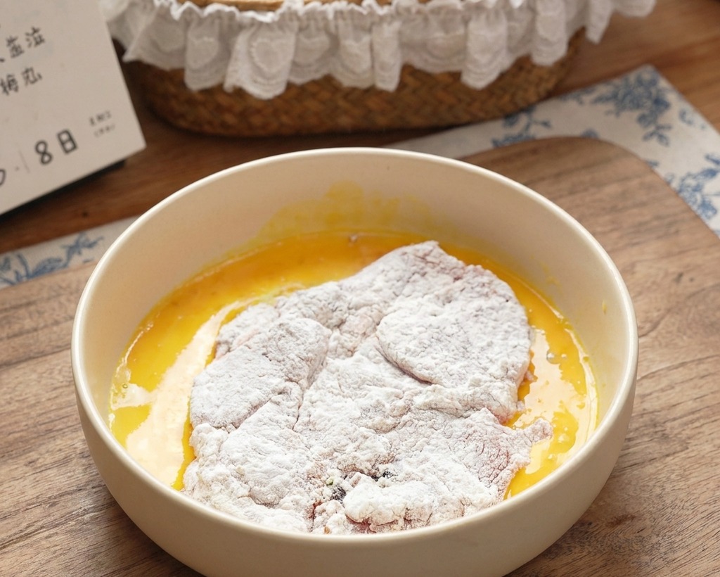 Ein mehliertes Schnitzel wird in eine Schüssel mit hellgelbem, verquirltem Ei getaucht.