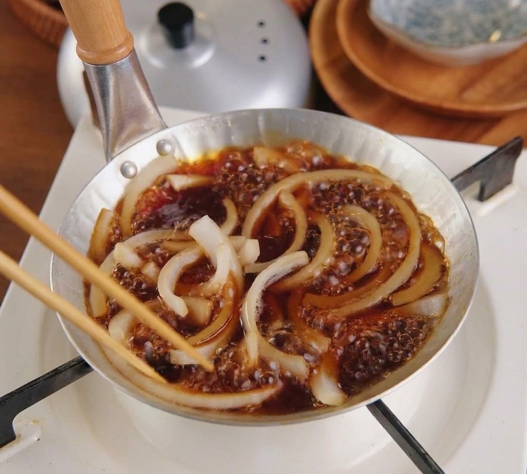 Fein geschnittene Zwiebeln köcheln in einer dunklen Sauce in einer silbernen Pfanne.