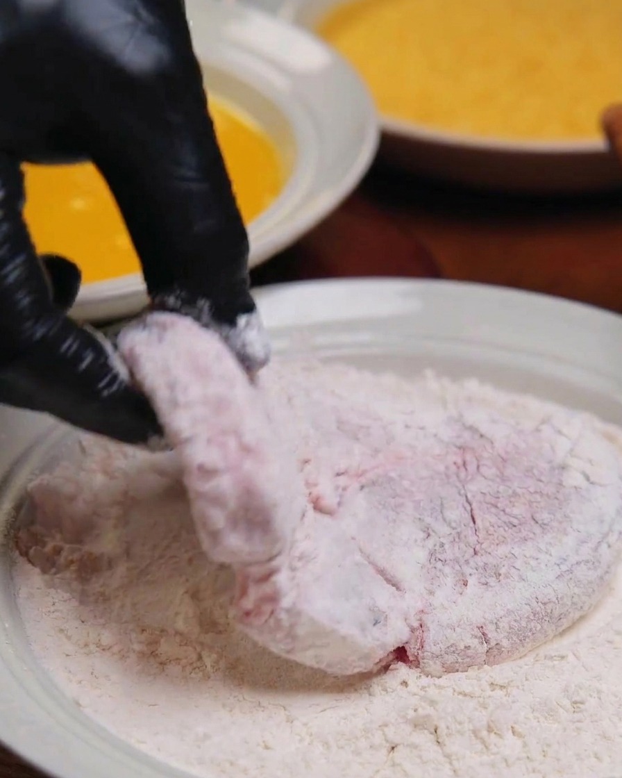 Eine Hand hält ein Stück Schweinefleisch, das vollständig mit weißem Mehl bestäubt ist, über eine flache Schale.