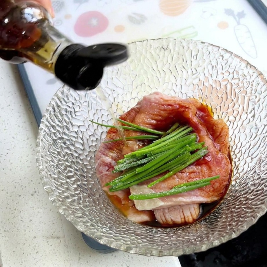 Eine Hand gießt Würzsauce über Schweinefleisch, Frühlingszwiebeln und weiße Stärke in einer Glasschüssel.
