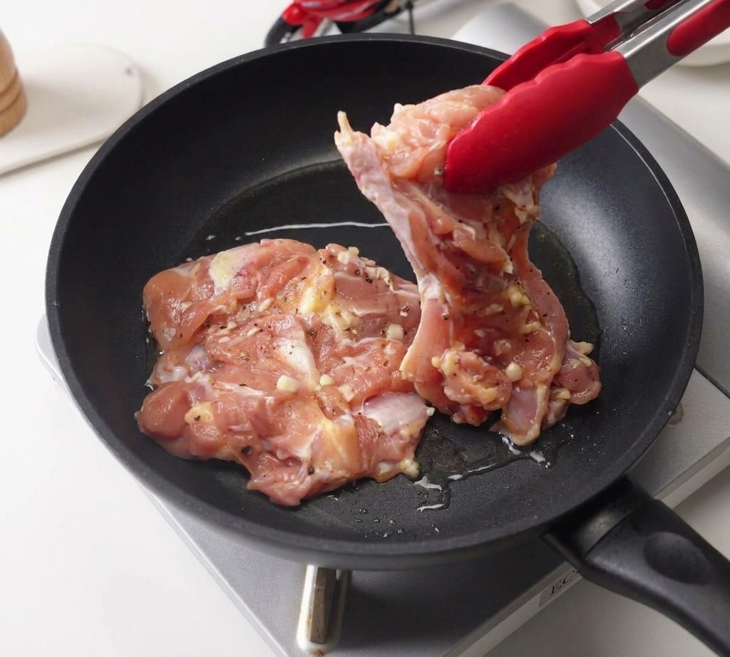 Eine Grillzange legt ein mariniertes Hähnchenstück mit der Hautseite nach unten in eine Pfanne mit Öl.