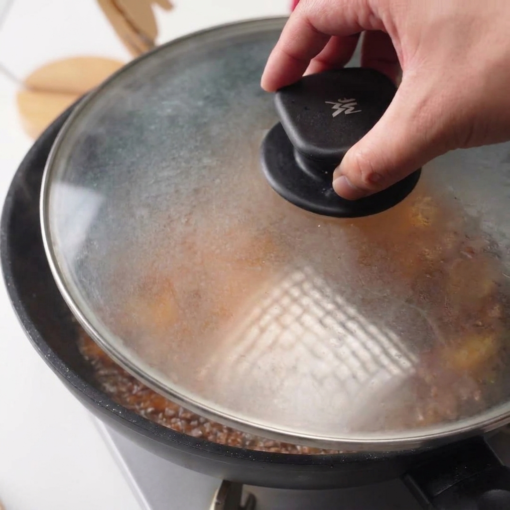 Ein Glasdeckel wird auf die Pfanne mit dem Hähnchen in köchelnder Sauce gesetzt.
