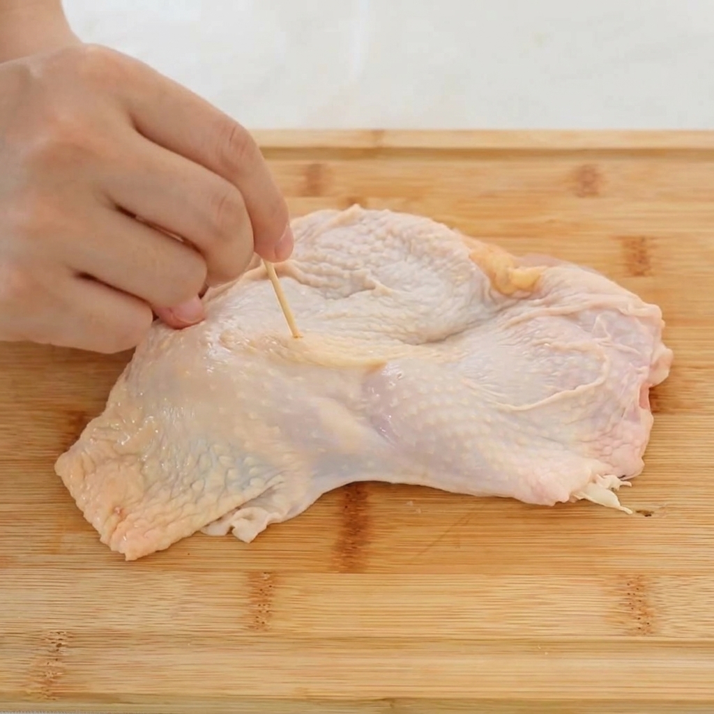 Nahaufnahme einer Hand, die mit einem Zahnstocher kleine Löcher in die Haut eines rohen Hähnchenschenkels sticht.