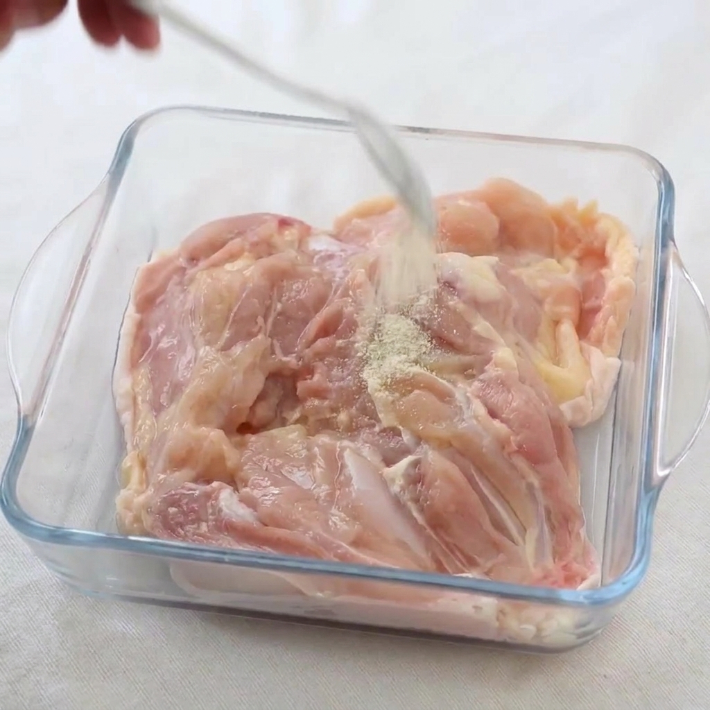 Weißer Pfeffer und Salz werden aus einem Löffel über rohe Hähnchenschenkel in einer Glasschale gestreut.