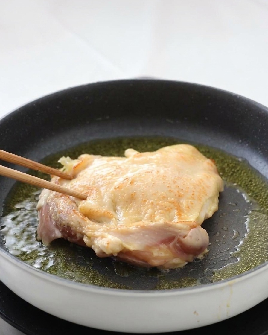 Ein Hähnchenschenkel wird mit Stäbchen in der Pfanne gewendet und offenbart eine perfekt gebräunte und knusprige Haut.