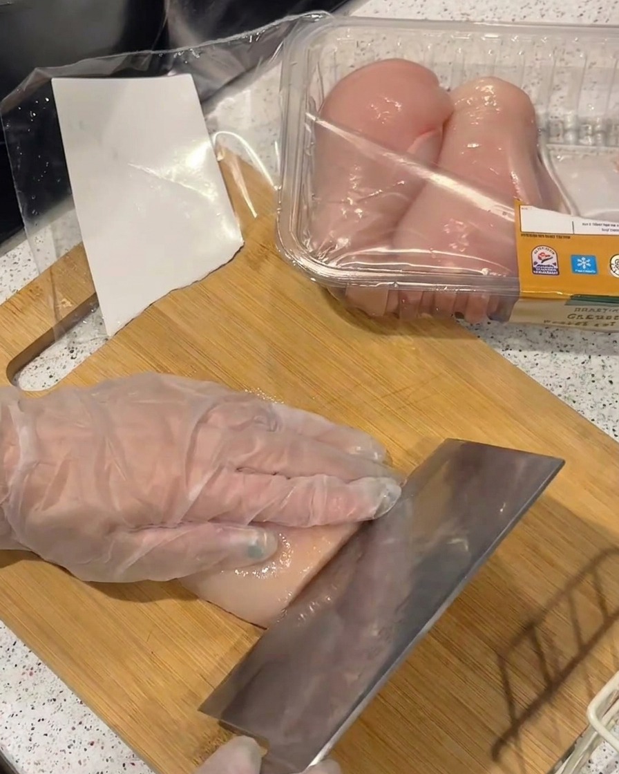 Eine behandschuhte Hand hält eine rohe Hähnchenbrust und schneidet sie mit einem großen Metallbeil in dünne Filets.