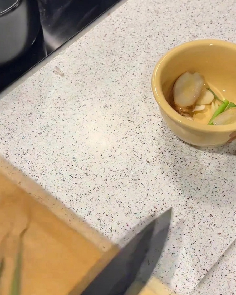 Eine gelbe Keramikschüssel mit frischen Ingwer- und Frühlingszwiebelscheiben auf einer Arbeitsplatte.