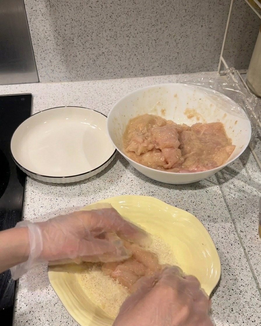 Ein mariniertes Hähnchenfilet wird fest in einen Teller mit hellem Panko-Paniermehl gedrückt.