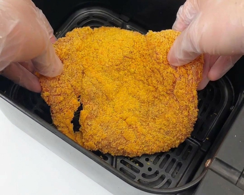Zwei Hände mit Handschuhen legen ein großes Stück paniertes Hähnchen in den schwarzen Korb einer Heißluftfritteuse.