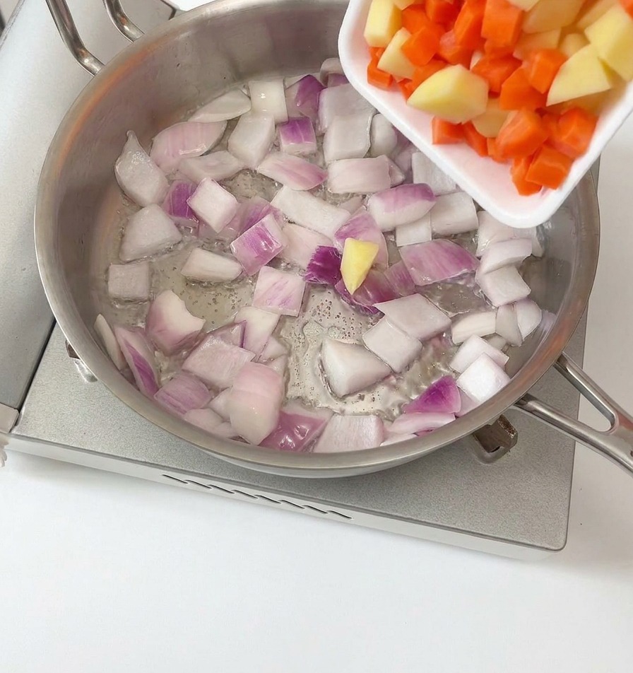 Gewürfelte Kartoffeln und Karotten werden aus einer weißen Schale in eine Edelstahlpfanne zu den gebratenen roten Zwiebeln gegeben.