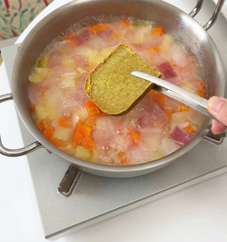 Eine Metallzange hält einen braunen Currywürfel über eine dampfende Pfanne mit Gemüse und Wasser.
