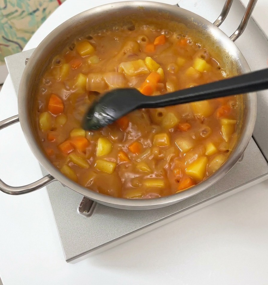 Ein schwarzer Pfannenwender rührt eine dicke, blubbernde braune Currysauce mit Kartoffel- und Karottenstückchen in einem silbernen Topf um.