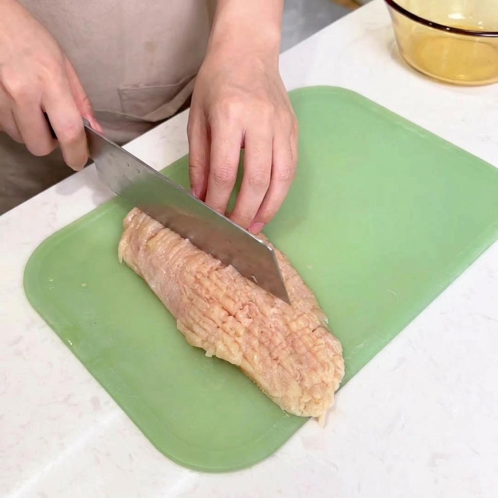 Der stumpfe Rücken eines großen Kochmessers schlägt auf ein flaches Stück rohes Hähnchen auf einem Schneidebrett.
