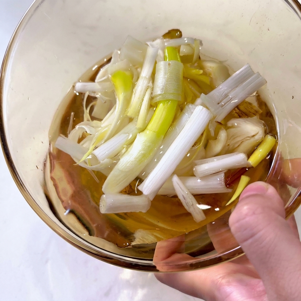 Nahaufnahme einer Glasschale mit klarer Flüssigkeit und Stücken von Frühlingszwiebeln sowie Ingwer-Scheiben.