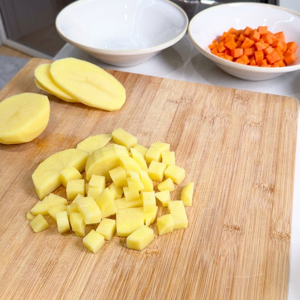 Gewürfelte Kartoffelstücke auf einem Holzbrett neben einer Schale mit Karottenwürfeln.