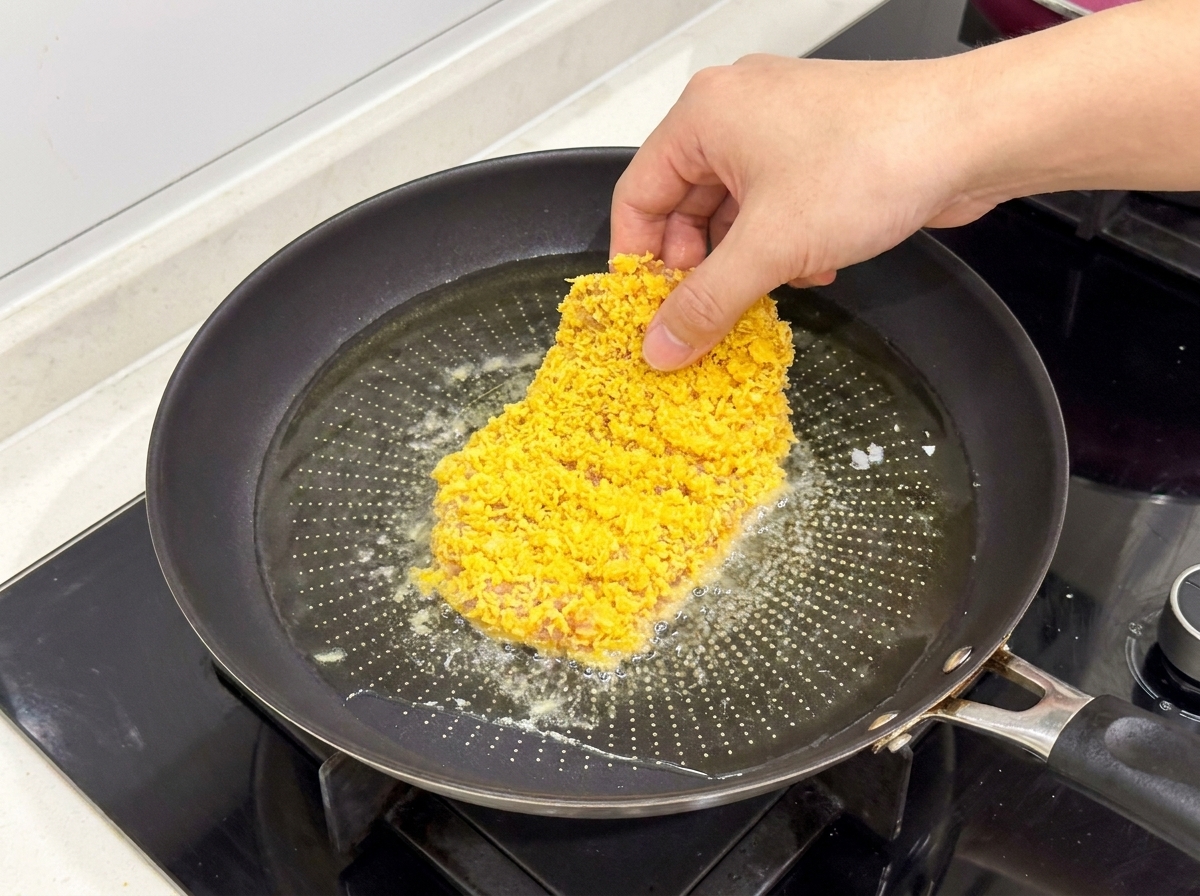 Ein paniertes Hähnchenschnitzel wird in eine Pfanne mit heißem, schimmerndem Öl gelegt.