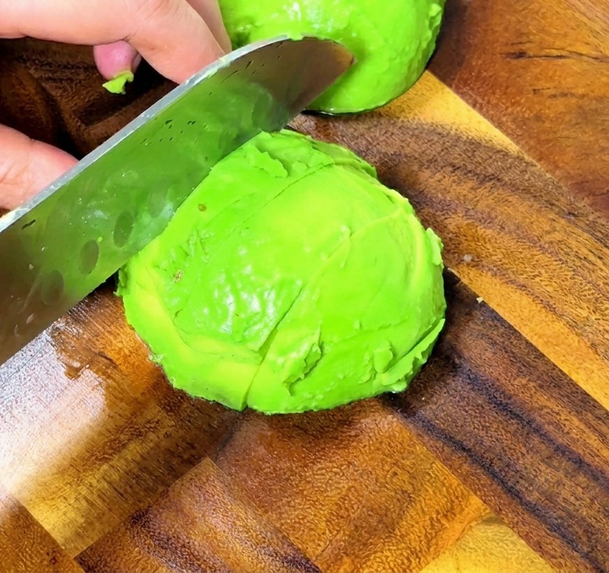 Nahaufnahme von Händen, die eine geschälte Avocado mit einem Profimesser in gleichmäßige Würfel auf einem Holzbrett schneiden.